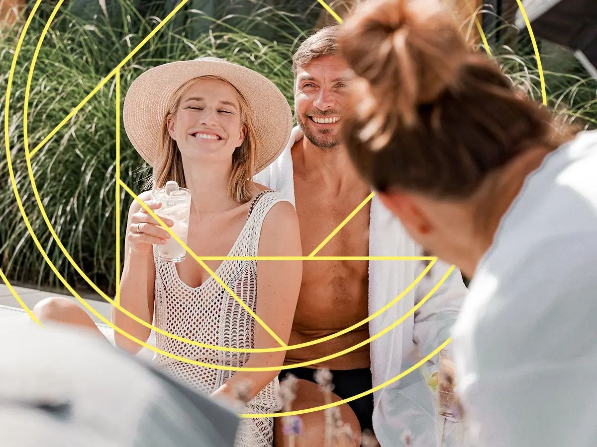 Paar sitzt lachend in sommerlicher Pool-Atmosphäre, während im Vordergrund fotografiert wird.
