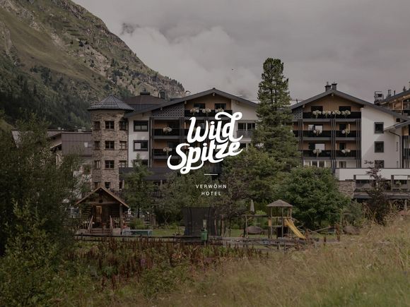 Außenansicht des Verwöhnhotels Wildspitze vor der alpinen Bergkulisse, mit platziertem Hotellogo im Vordergrund.